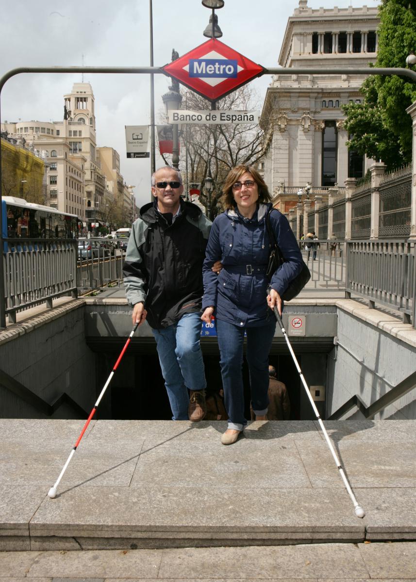 Una persona con bastón rojo-blanco pasea junto a otra persona con bastón blanco